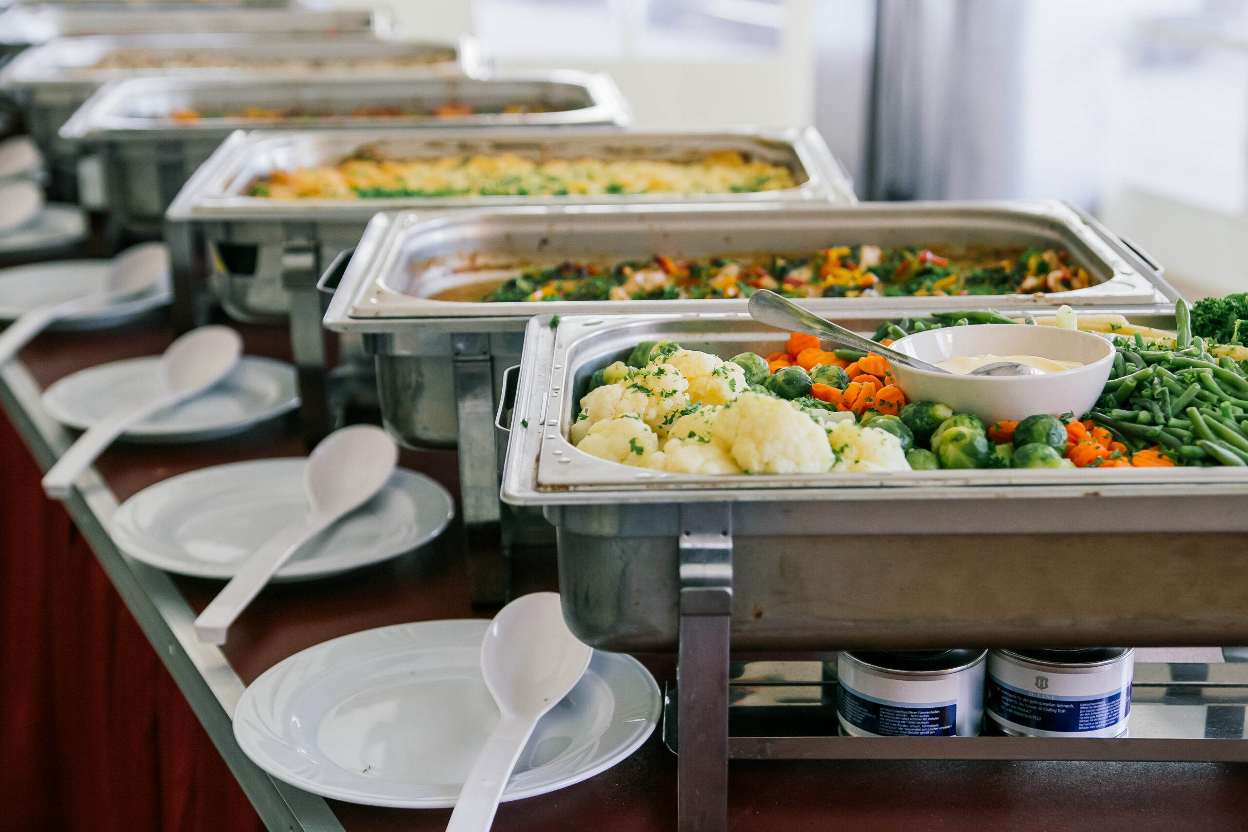 A buffet line featuring several silver chafing dishes filled with various hot dishes, including vegetables like green beans, brussels sprouts, carrots, and cauliflower, alongside white plates and serving utensils. A buffet line featuring several silver chafing dishes filled with various hot dishes, including vegetables like green beans, brussels sprouts, carrots, and cauliflower, alongside white plates and serving utensils.