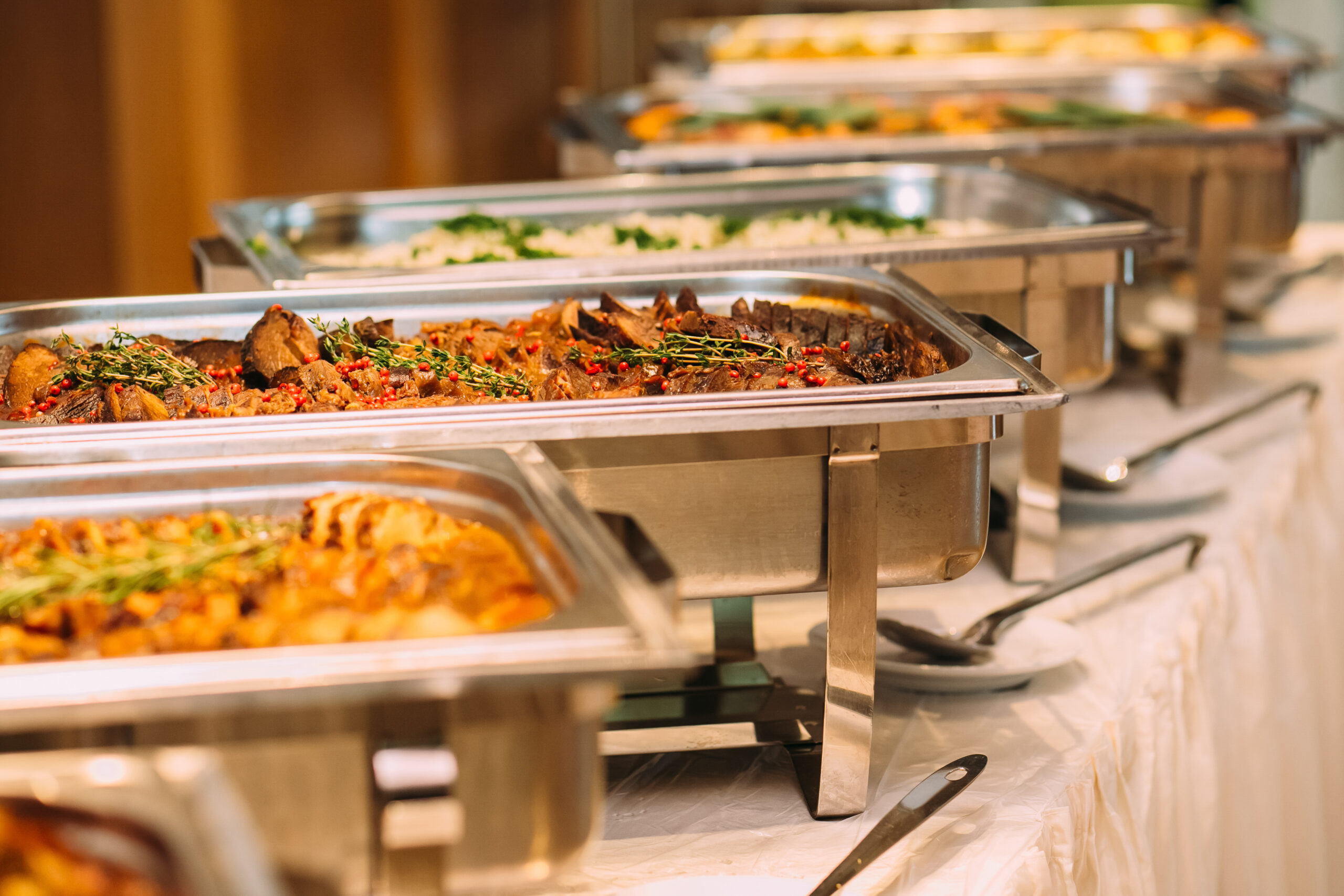 Catering Food Wedding Table A line of stainless steel chafing dishes filled with various hot dishes, including what appears to be a meat dish garnished with herbs, on a white tablecloth. A line of stainless steel chafing dishes filled with various hot dishes, including what appears to be a meat dish garnished with herbs, on a white tablecloth.