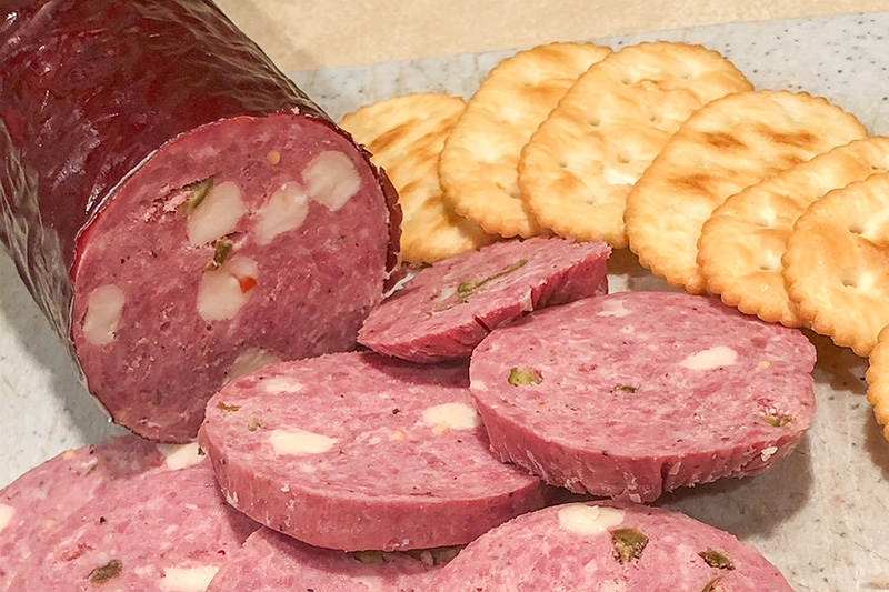 A close-up of a large log of jalapeno and cheese salami, partially sliced to reveal chunks of white cheese and green jalapenos within the reddish meat, served alongside several golden crackers on a light-colored surface. A close-up of a large log of jalapeno and cheese salami, partially sliced to reveal chunks of white cheese and green jalapenos within the reddish meat, served alongside several golden crackers on a light-colored surface.