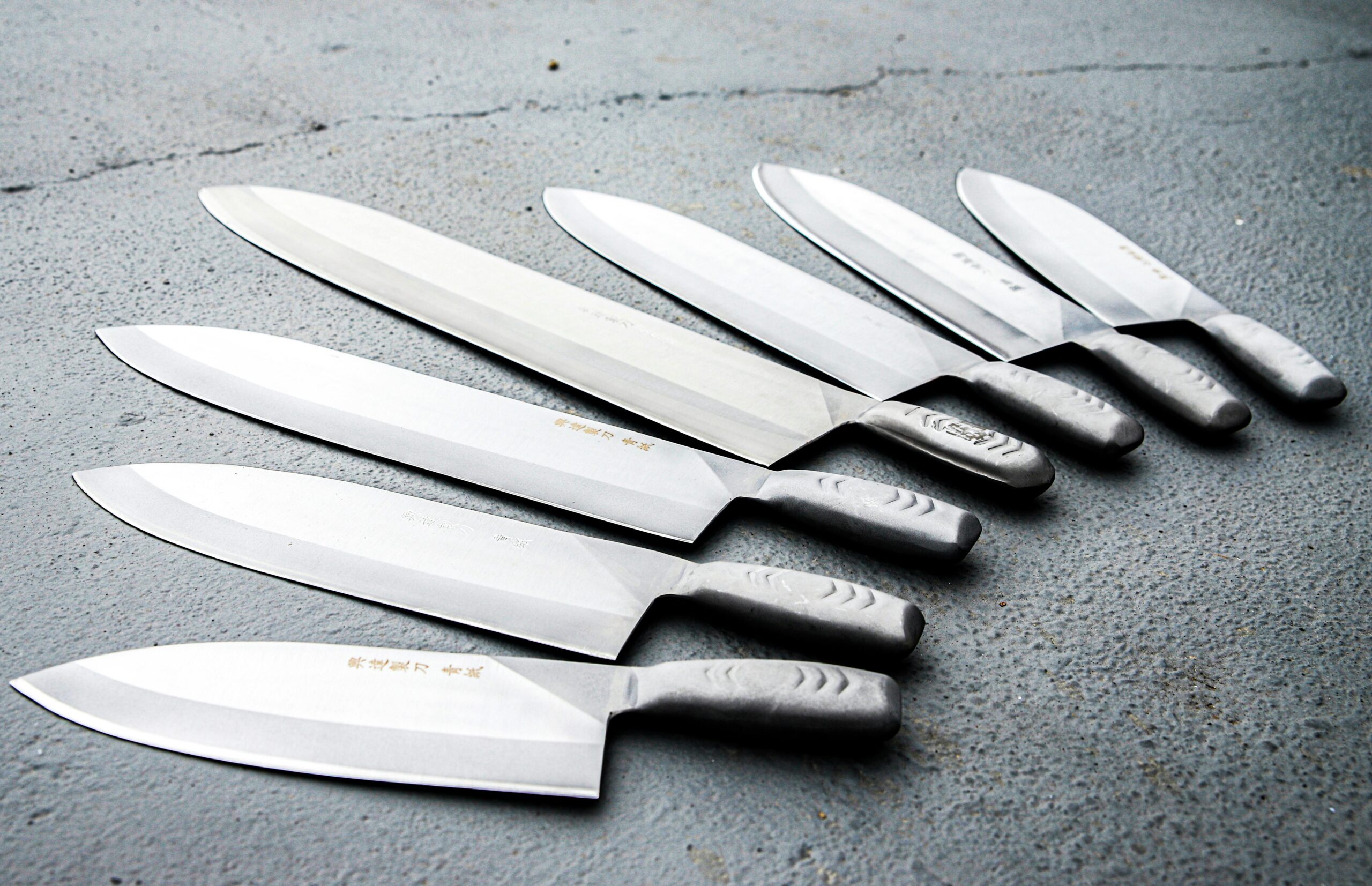 A set of six professional kitchen knives, with silver blades and grey textured handles, are neatly arranged in two rows on a dark, textured surface. A set of six professional kitchen knives, with silver blades and grey textured handles, are neatly arranged in two rows on a dark, textured surface.