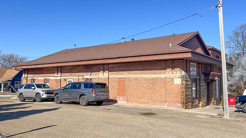 The exterior of Ruzicka's Meat Market, a brick building with a brown roof, stands under a clear blue sky. Two SUVs are parked in front, and a plume of white smoke rises from the right side of the building. The exterior of Ruzicka's Meat Market, a brick building with a brown roof, stands under a clear blue sky. Two SUVs are parked in front.
